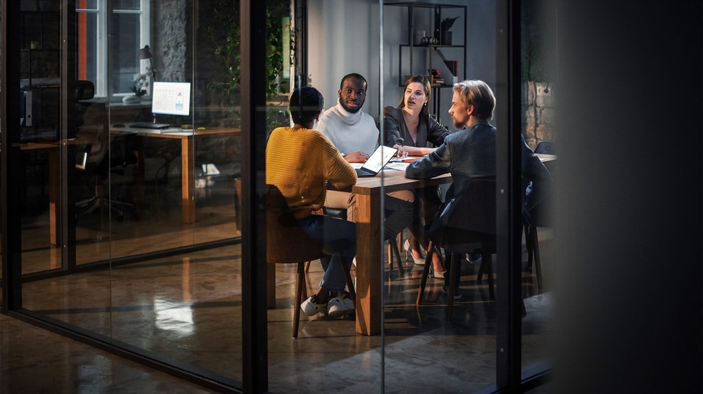 A group of people sit around a wooden table in a modern glass‑walled meeting room, reviewing work on laptops and documents.