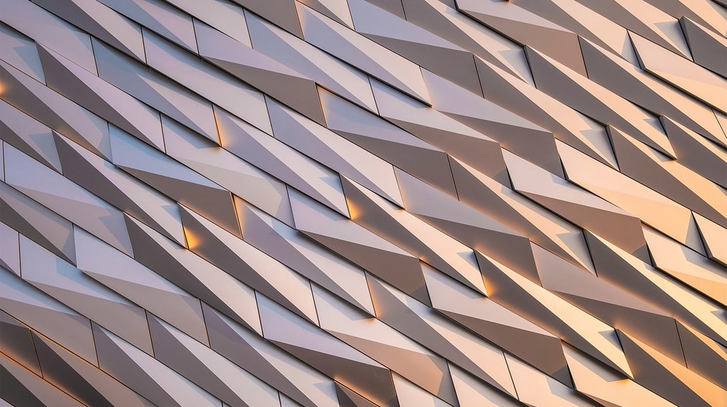An abstract architectural façade made of angled metallic panels that catch soft light and create a repeating geometric pattern.
