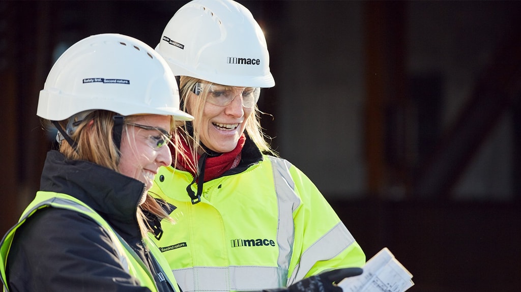 Two construction workers in hard hats and high-visibility jackets are reviewing plans on a clipboard at a construction site.