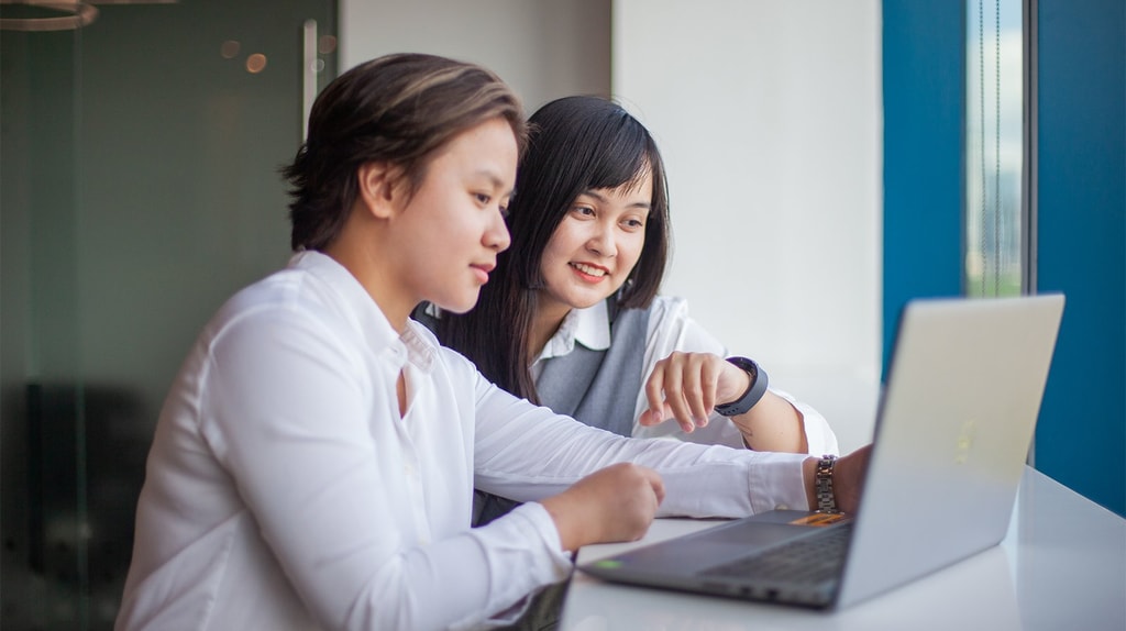 Two people sit at a desk by a window, working together on a laptop as one person points toward the screen.