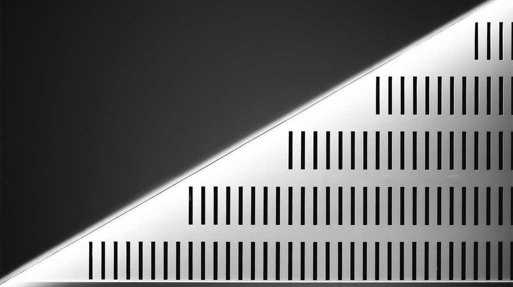 A minimalist black‑and‑white architectural detail shows diagonal lighting cutting across a wall of evenly spaced vertical slits.