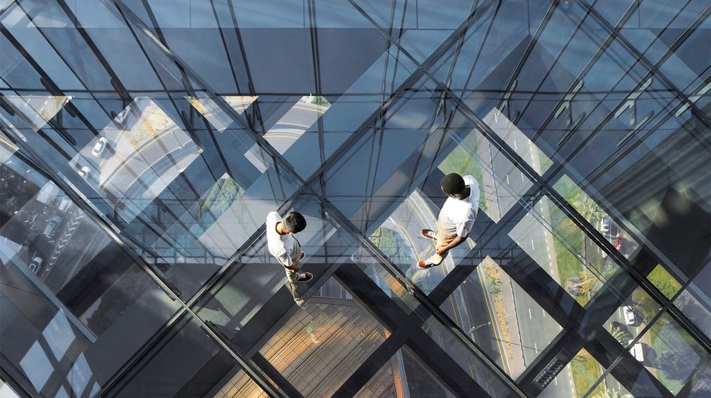 Two people walk across a transparent glass floor in a high‑rise building, with reflections revealing the city streets and structures below.