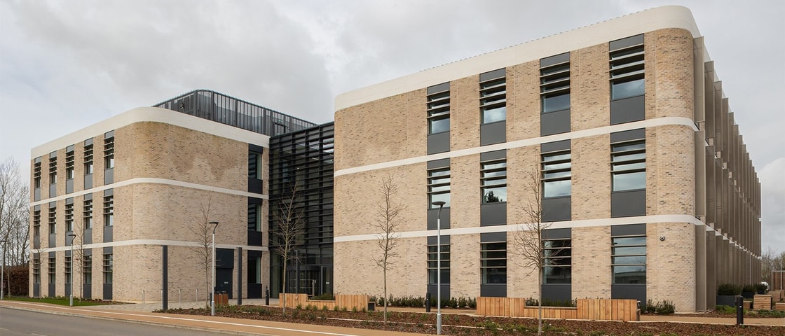 Modern commercial or institutional building with light brickwork, large vertical windows, and horizontal black louvers. The structure features rounded corners, white horizontal bands between floors, and a landscaped frontage with young trees and wooden fencing under an overcast sky.