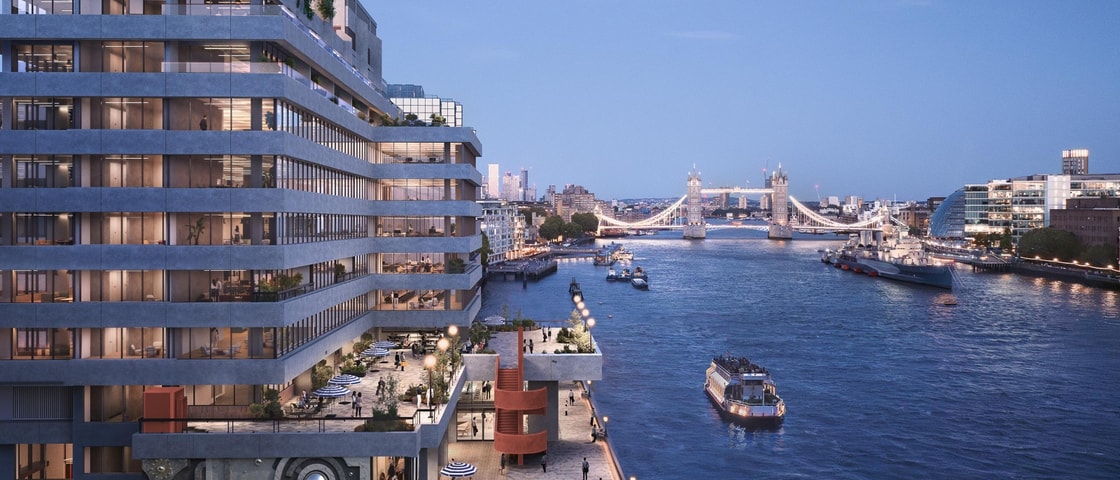 A CGI architectural rendering of a modern riverside building overlooking the River Thames at dusk. The multi-storey development features large glazed windows, stepped terraces, rooftop planting and active waterfront spaces with outdoor seating and pedestrians. Tower Bridge is illuminated in the background, while several boats travel along the river, framed by the London skyline under a blue evening sky.