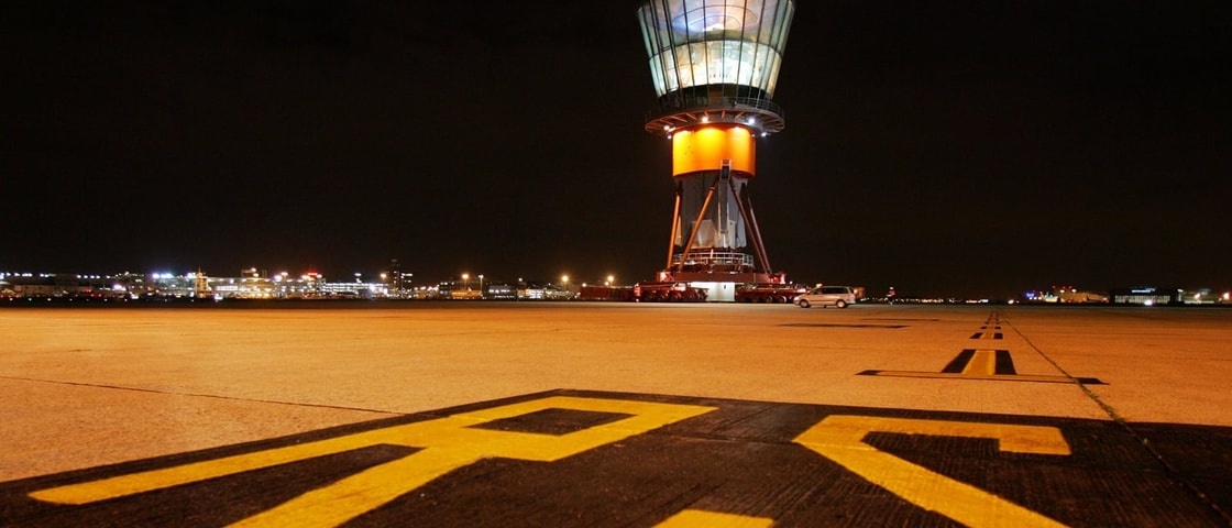 Night-time view of an airport runway with large yellow runway markings in the foreground. In the distance, an illuminated air traffic control tower sits on a large transport platform, with airport buildings and lights stretching across the horizon beneath a dark sky. The low-angle perspective emphasises the scale of the runway and the tower