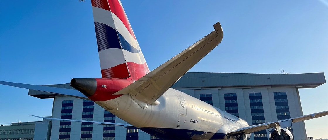 A British Airways airplane's tail in the foreground, with a hangar behind and another plane flying in the clear blue sky above.