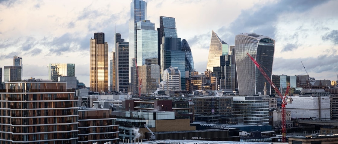 Panoramic view of the City of London skyline featuring modern glass skyscrapers, including the Gherkin, the Leadenhall Building and 20 Fenchurch Street (the Walkie Talkie). Residential and commercial buildings fill the foreground, while several red construction cranes are visible among the high-rise towers.