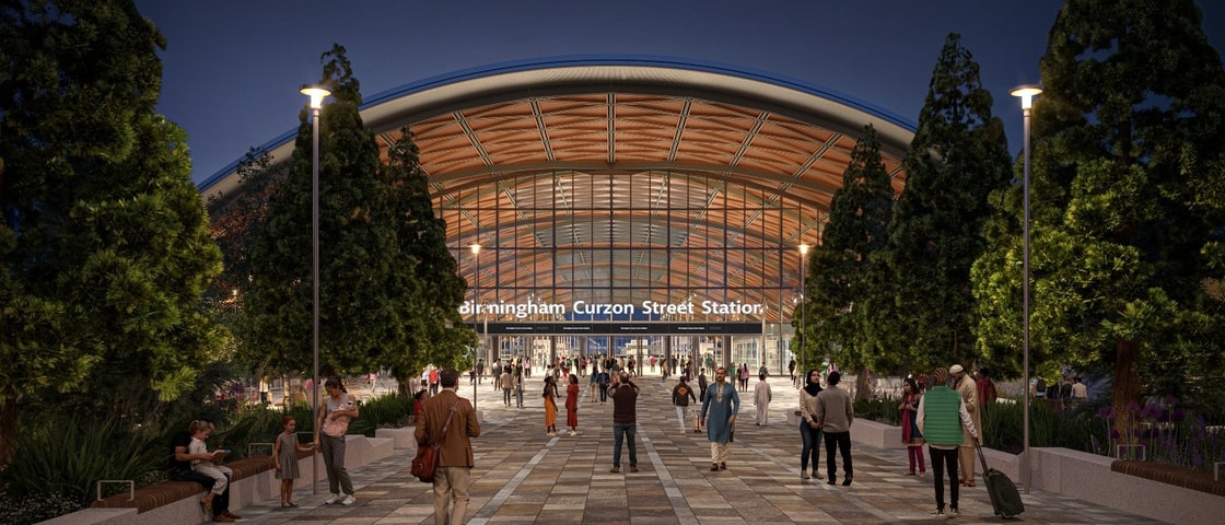 Profile view of the HS2 Curzon Street station’s exterior, showing the sweeping curved roof with timber-lined structure, large glass façade, and surrounding landscaped public spaces under a bright sky.