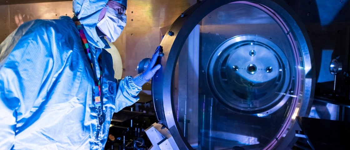 A person wearing full cleanroom protective clothing and face covering stands beside a large circular scientific instrument, looking through its transparent chamber. The scene is lit with blue and purple tones, and the equipment appears to be part of a high‑tech laboratory or manufacturing environment.