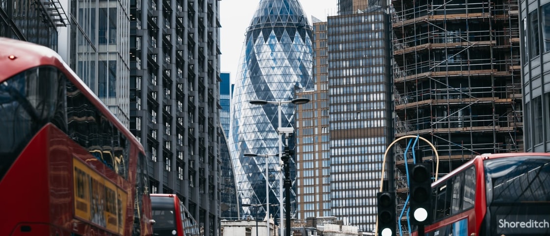 View along a busy London street lined with tall modern buildings, with the Gherkin skyscraper centred in the background. Red double-decker buses travel in both directions in the foreground, while a building on the right is covered in scaffolding. 