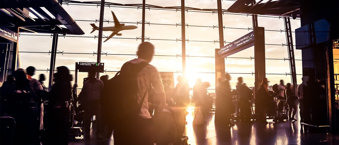 Silhouetted travelers move through a bright airport terminal with large glass windows as a plane flies past in the sunset outside.
