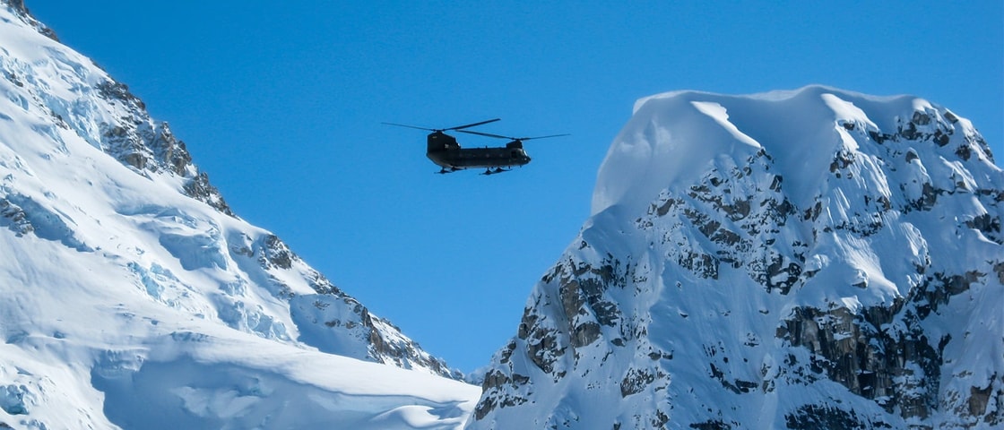Helicopter flying between snow-covered mountain peaks under a clear blue sky.
