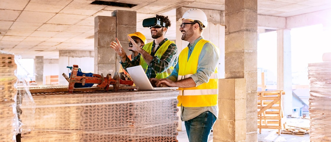 Two construction workers in safety vests collaborate on a laptop amidst a partially built structure, discussing plans and using technology.
