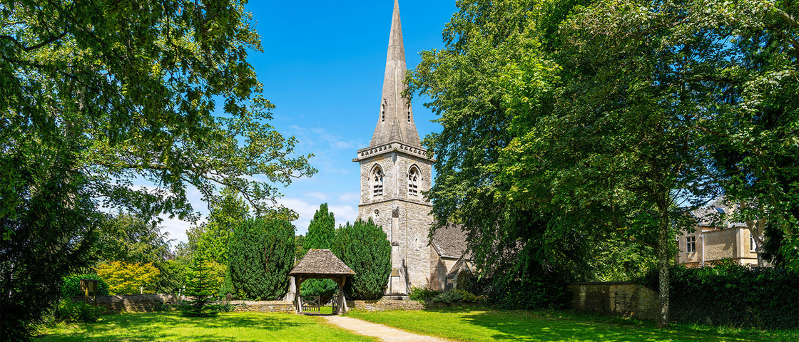 A stone church with a tall spire stands at the end of a grassy pathway, surrounded by large trees and bright greenery under a clear blue sky.