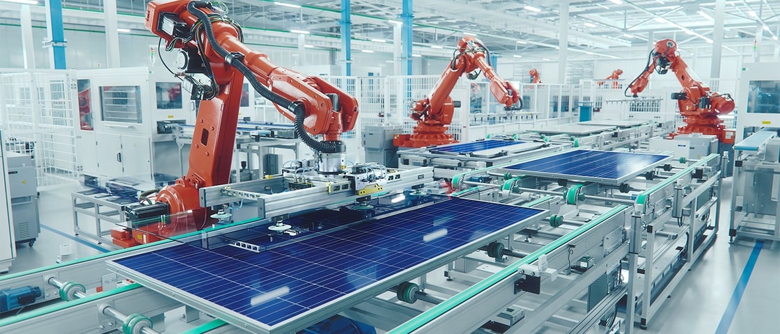 A row of orange robotic arms assembles large blue solar panels on an automated production line inside a bright, modern manufacturing facility.