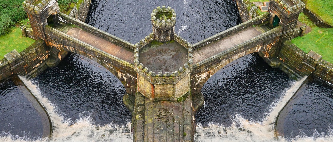 Aerial view of a historic stone dam spillway with symmetrical walkways and turrets, directing water through multiple channels surrounded by green landscape.