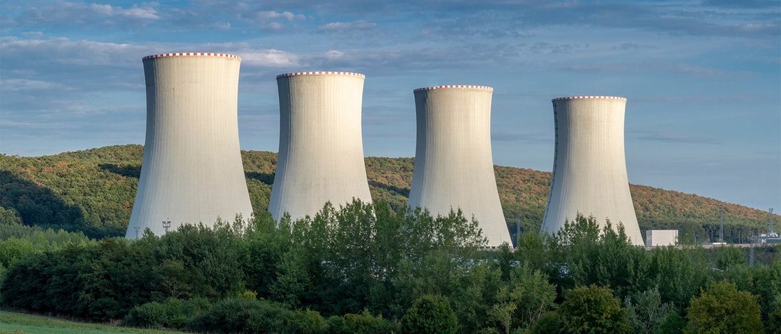 Four large cooling towers of a nuclear power plant rise behind a green forested area with hills in the background under a partly cloudy sky.