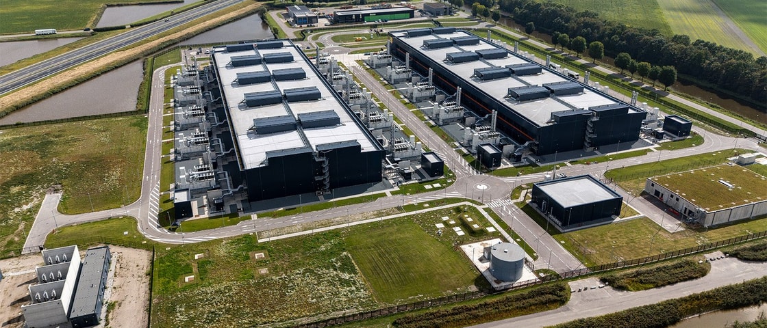 An aerial view of a large industrial facility with multiple long rectangular buildings, service roads, and surrounding fields in a rural landscape.