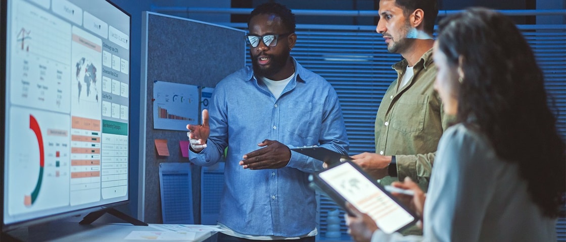 A group of people in an office setting review data visualizations on a large screen while discussing analytics and using a digital tablet.