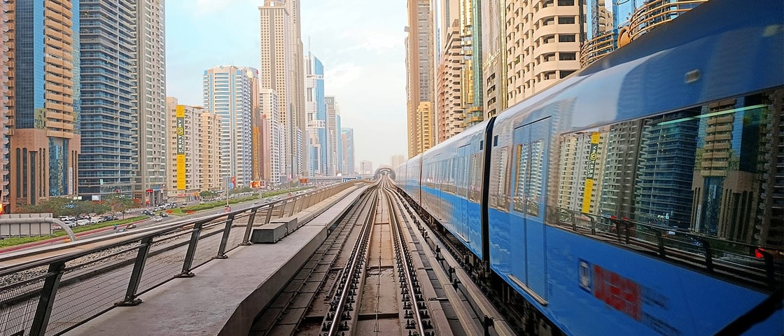 Blue metro train travelling along elevated tracks through a dense modern cityscape of high-rise buildings, viewed from a central track perspective under a station canopy.