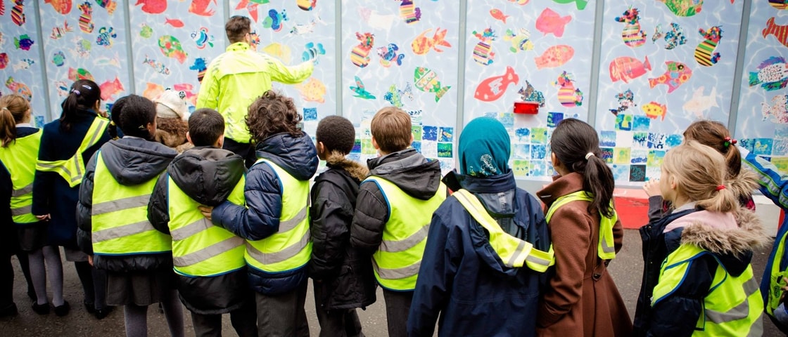A group of children in high‑visibility vests stand in front of a colourful wall mural featuring fish artwork as an adult points out details.
