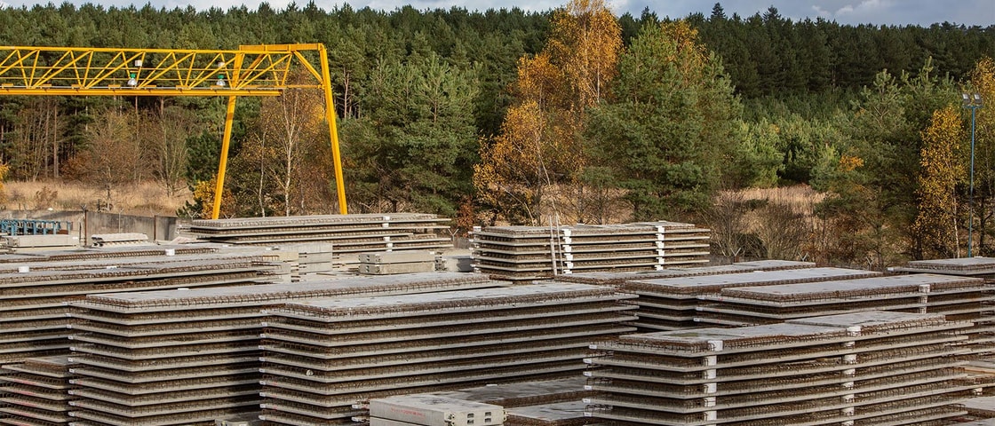 A construction site with stacked concrete slabs, under a yellow crane, surrounded by lush greenery and a cloudy sky.