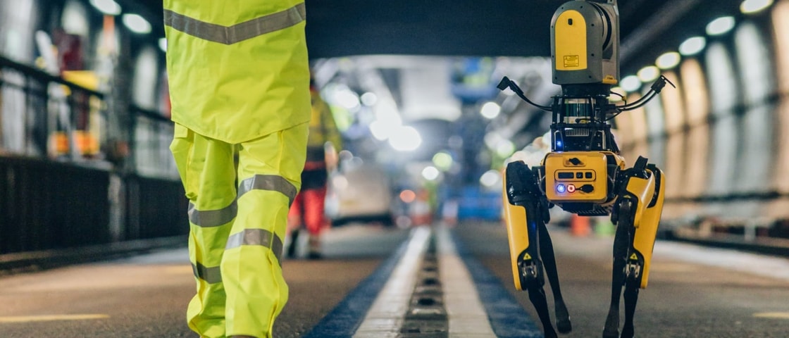 A worker in high-visibility yellow protective clothing walks alongside a four-legged robotic scanning device on a railway platform or track inspection area. The robot, equipped with sensors and cameras, moves along the platform edge under bright station lighting, with rail infrastructure blurred in the background.