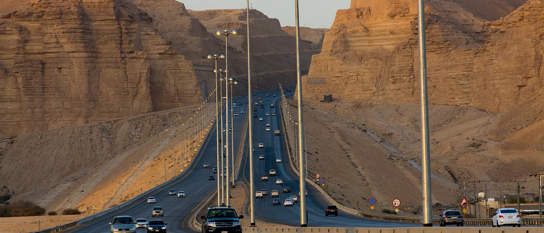 A multi-lane highway carrying steady traffic winds through a dramatic desert landscape, with tall streetlights lining the road as it passes between towering sandstone cliffs under warm evening light.