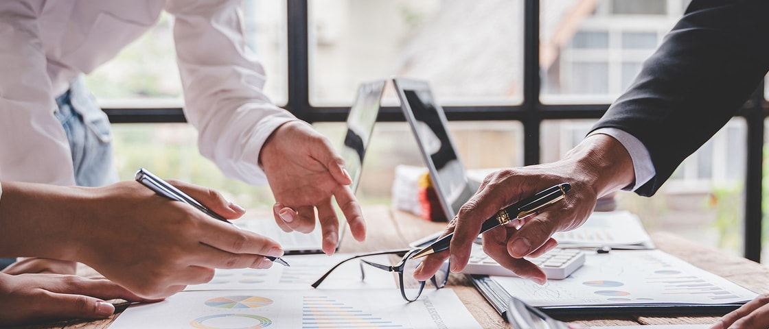 Two professionals discussing charts and reports at a wooden table, with a laptop and glasses nearby, in a bright office setting.