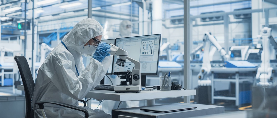 A person in full protective laboratory gear working with a microscope at a workstation equipped with a computer inside a high-tech industrial lab environment.