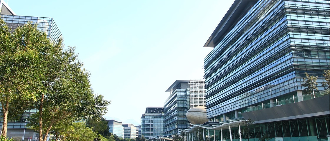 A collection of contemporary glass office buildings lining a landscaped waterfront, with trees, reflective waterways and modern architecture creating a business district setting.