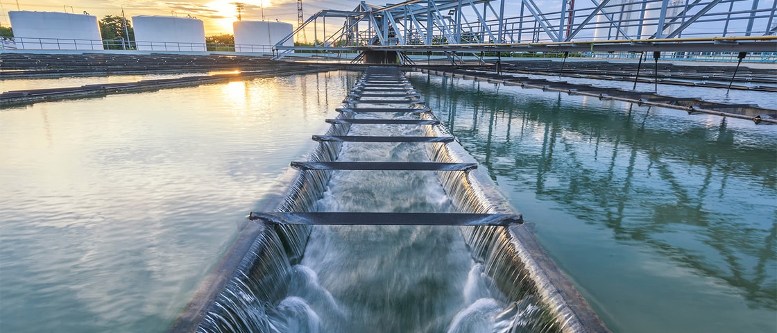 A wide view of a water treatment facility showing flowing channels, metal walkways and settling basins under early evening light.