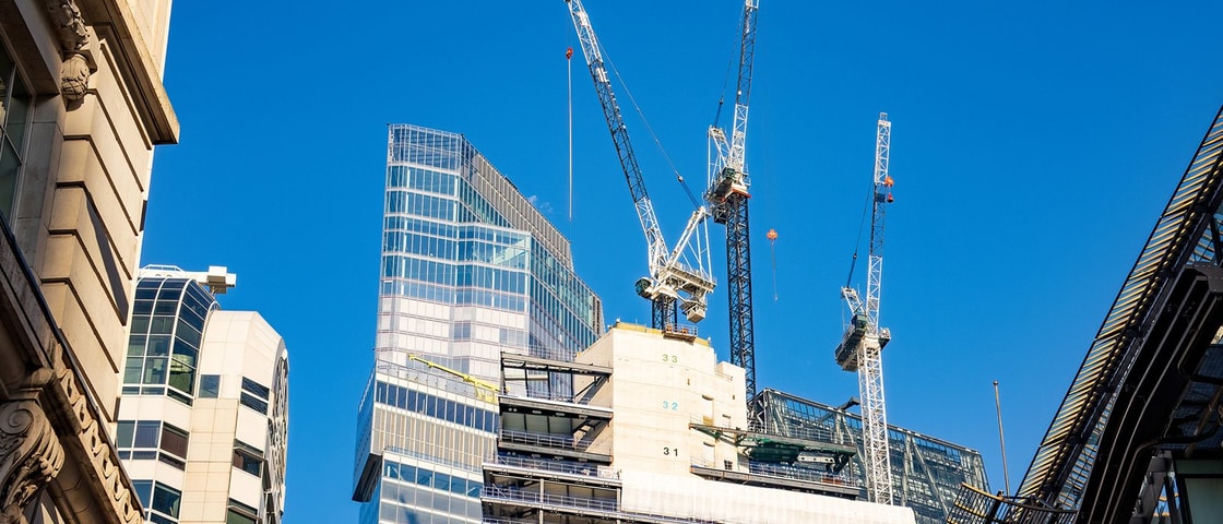 A construction site with cranes and partially built high‑rise structures stands against a clear blue sky in a modern urban area.