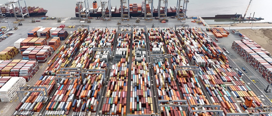 An aerial view of a large container port showing rows of multicoloured shipping containers stacked in long grids, with cranes lining the quay and cargo ships docked beside the water.