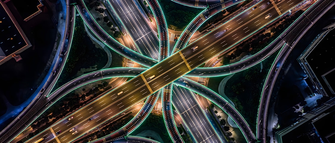 An aerial view of a brightly illuminated motorway interchange at night shows intersecting highways, flowing traffic, and illuminated roadways forming a complex transport network.