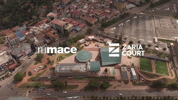 An aerial view of a modern urban complex featuring multiple green-roofed buildings, sports courts, parking areas, and surrounding roads with dense city structures in the background.