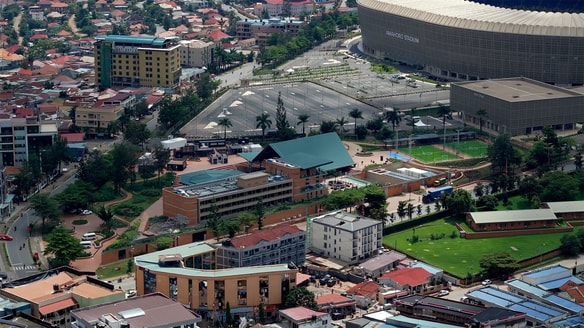 Aerial view of a city district featuring the large circular facade of a stadium, surrounding parking lots, green sports fields, and clusters of modern buildings with red-tiled roofs.