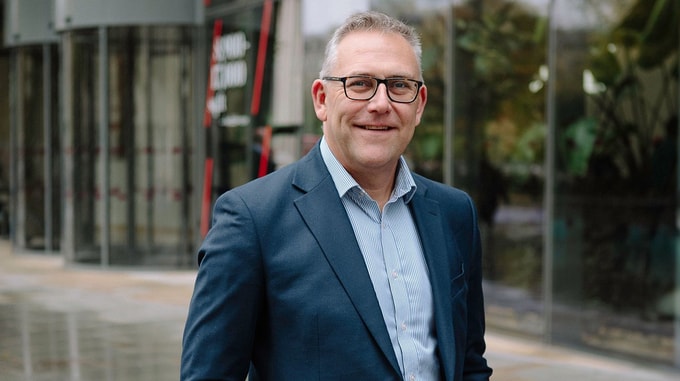 A person wearing glasses and a suit jacket stands outdoors in front of a modern glass building.