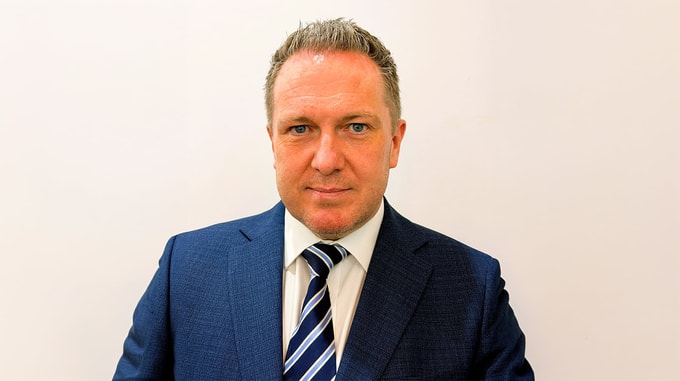Professional headshot of Christopher McMahon wearing a blue suit jacket, white shirt and striped tie, photographed against a plain white background.
