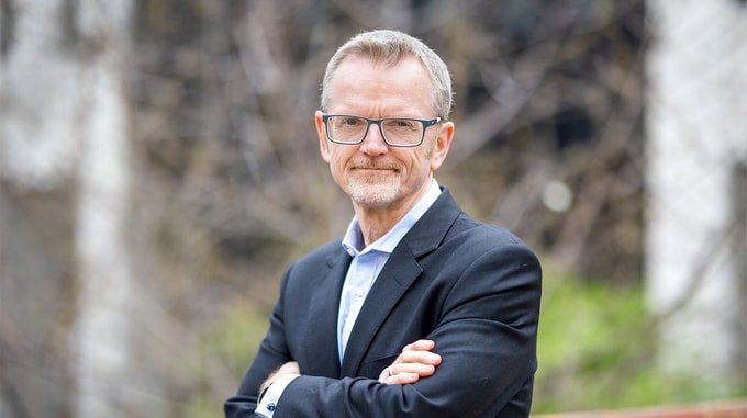A person wearing glasses and a dark blazer stands outdoors with arms folded, positioned in front of a softly blurred background of trees and greenery.