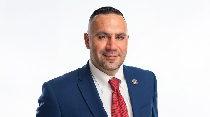A person wearing a blue suit jacket, white dress shirt, and red tie is posed against a plain white background.