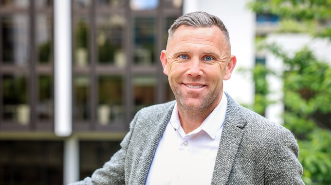 Portrait headshot of an adult with short hair wearing a grey textured blazer and light shirt, standing outdoors in front of a blurred modern building and greenery.