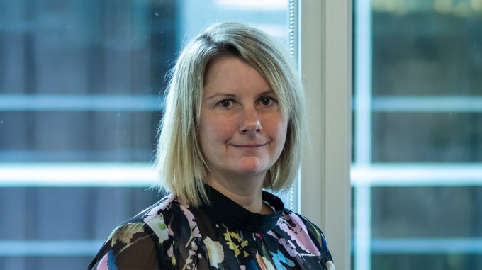 Professional headshot of an individual with shoulder-length light hair wearing a patterned blouse, standing indoors by a window with soft natural light and a blurred office building visible in the background.