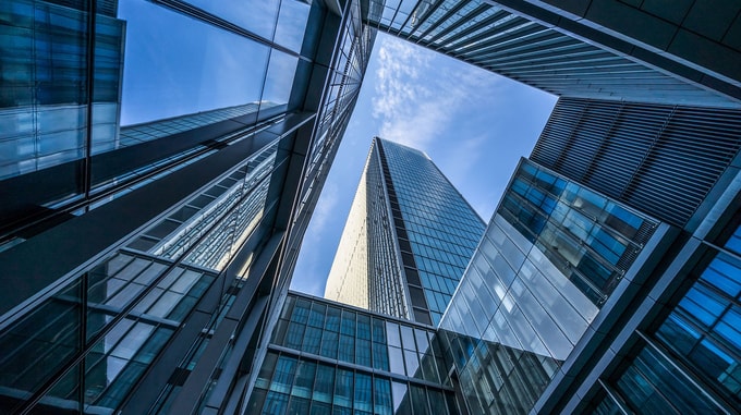 A tall glass skyscraper is framed by surrounding reflective buildings, viewed from below against a bright blue sky.
