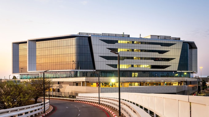 A modern glass-fronted office building with sleek geometric design and reflective windows, viewed from a curved roadway at sunset.