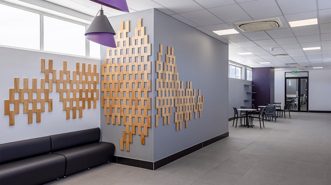 A modern office lounge with geometric wooden wall art, black seating, and tables arranged for informal meetings or relaxation.