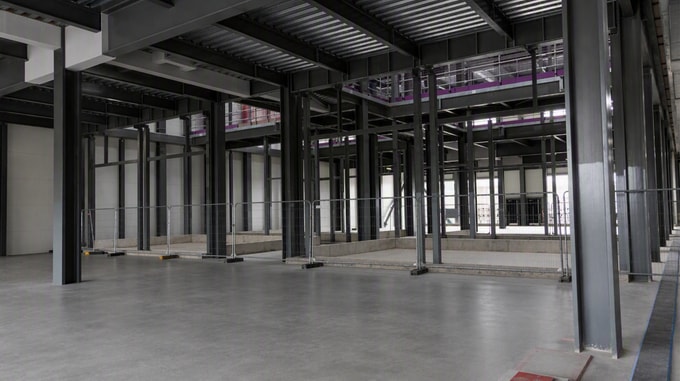 Interior view of a large industrial facility under construction, featuring black steel columns and beams, a concrete floor, and a partially visible red mobile access platform positioned behind a structural column.