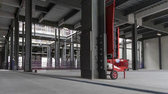 Wide view of an industrial building interior under construction, with exposed steel framing, temporary safety fencing, concrete foundations, and open floor space within the partially completed structure.