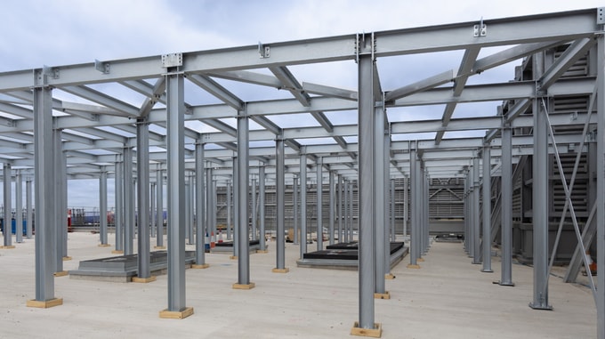 Steel framework under construction on an outdoor rooftop or plant deck area, showing a grid of grey steel columns and beams installed on a concrete surface beneath an overcast sky.