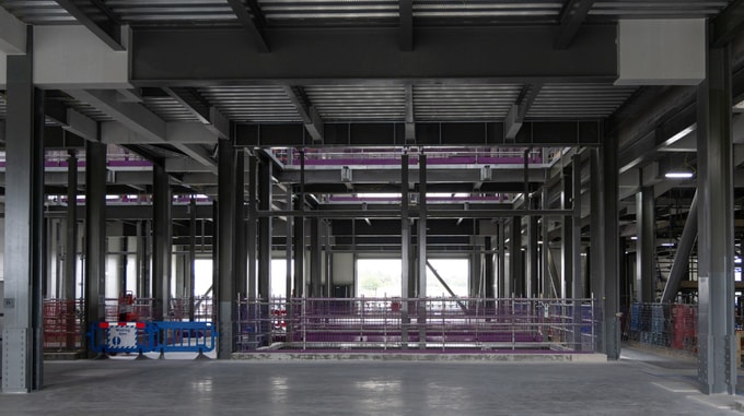 Interior view of a partially completed steel-framed building, featuring structural columns and beams, safety fencing around work areas, and an open concrete floor.